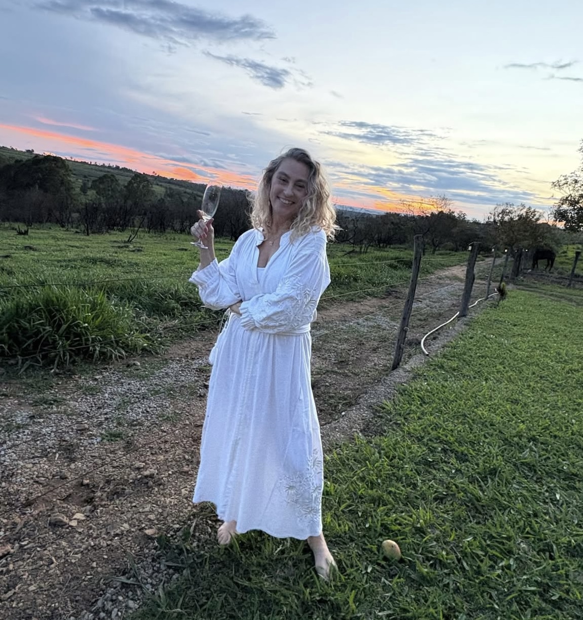 Mulher de vestido claro em varanda do Spa do Vinho, taça de vinho na mão, contemplando os vinhedos ao entardecer